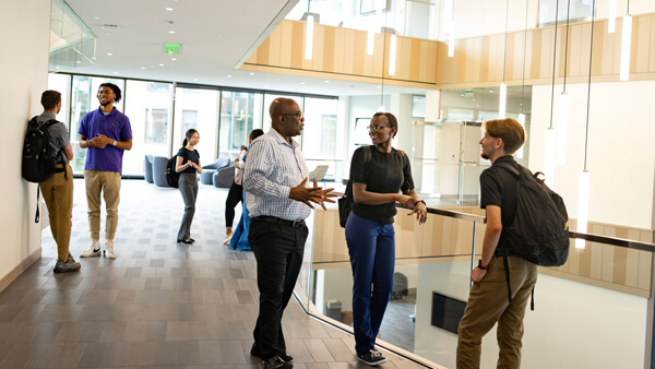 Students and professors talk by a glass balcony wall of a brightly lit, three-story atrium