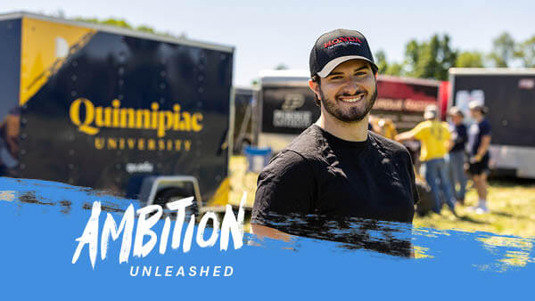Alex Petrarca stands outside a trailer reading Quinnipiac University used to transport the Quinnipiac baja vehicle