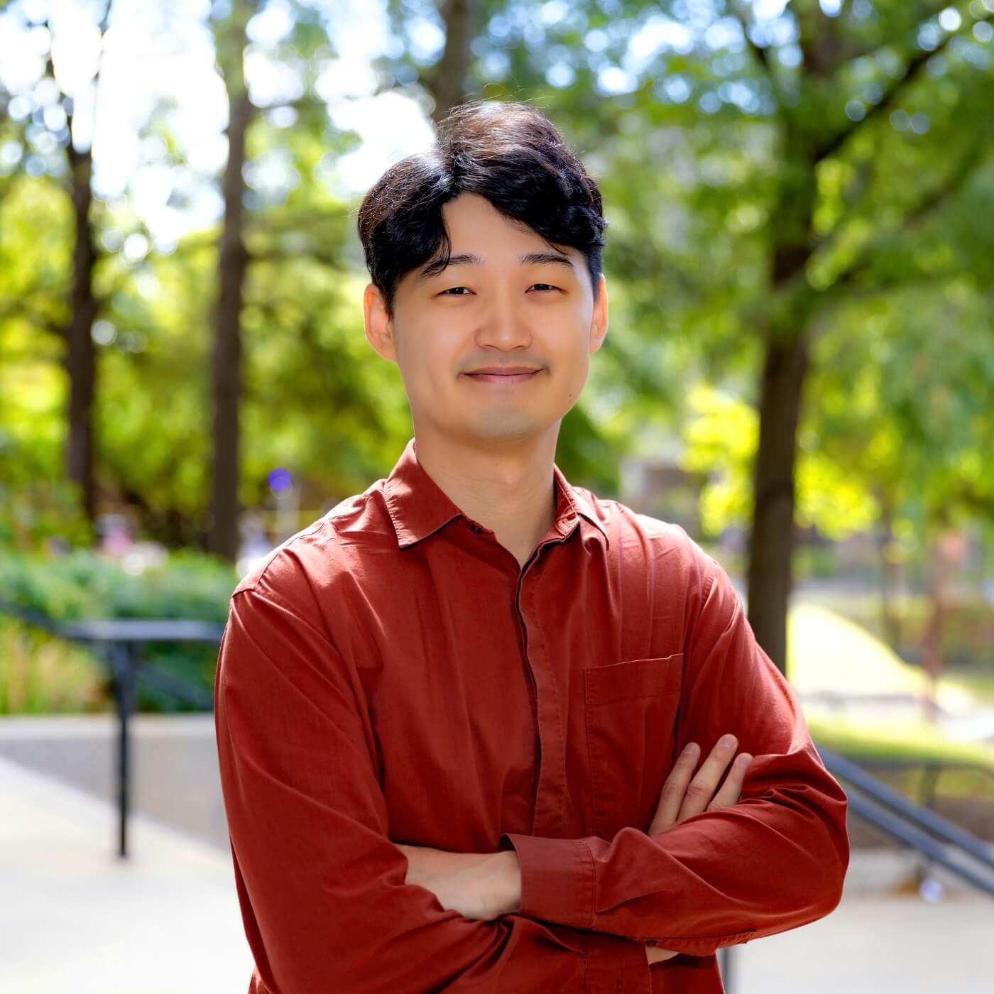 Deokkyung Ock smiling with his arms crossed against some green trees