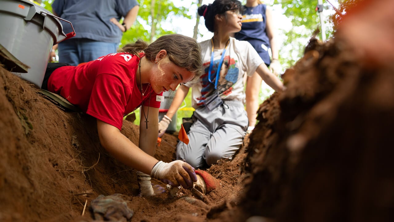 A high school student carefully excavates soil with a small trowel during a forensic science activity, uncovering a staged human skull wearing a red beanie as part of a mock crime scene at Quinnipiac University.