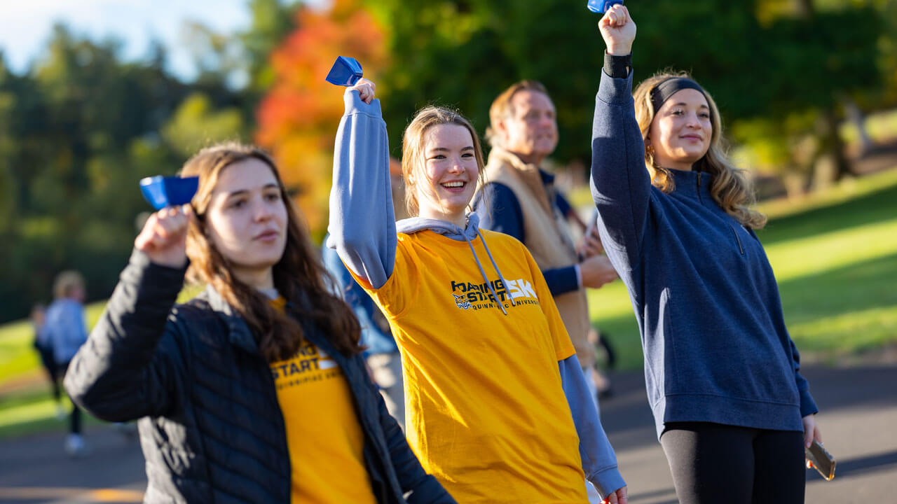 Three student volunteers cheering on the runners with cowbells
