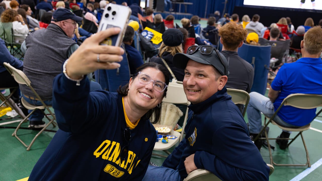 Two individuals pose for a selfie amongst a crowd of people sitting in chairs
