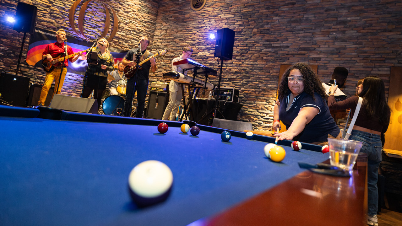 A female student playing pool with a band playing in the background