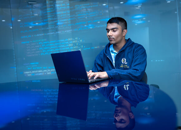 A male student seated at a table works on a laptop in a workspace with lines of code projected on the walls.