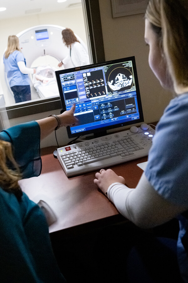 Student viewing imaging results on a computer while a patient goes through on MRI machine
