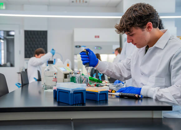 A male student wearing blue gloves uses a pipette dropper and other lab equipment.