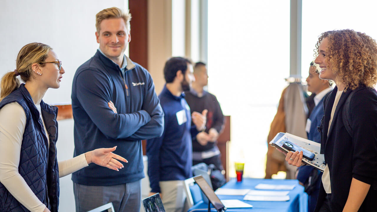 An exhibitor talks over a table to a fair attendee holding a stack of papers.