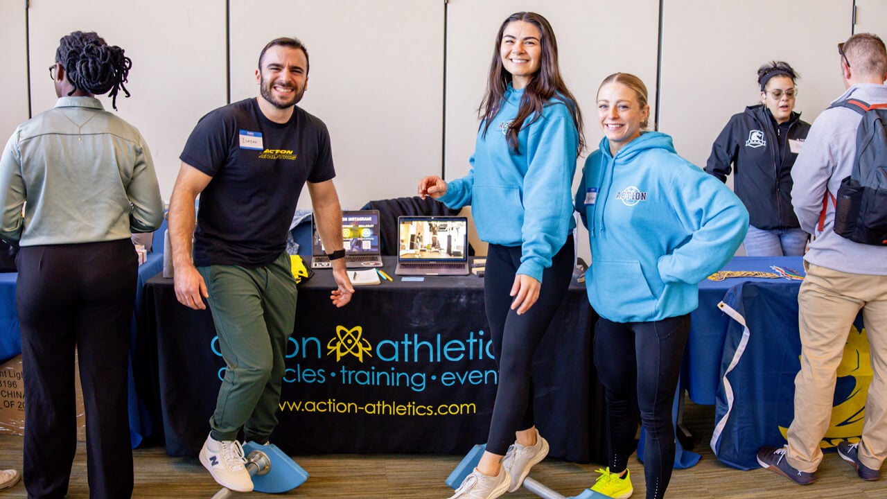 Three students in athletic wear pose in front of a booth showcasing their athletic training business.
