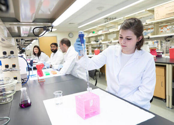 A woman sits doing medical lab work at a counter with 2 men and a woman in the background.