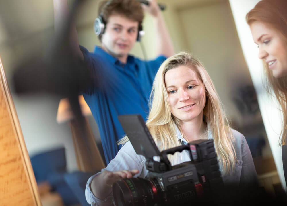 Ashley Brandon showing a female student something on a video camera while a male student in the background holds a microphone boom.