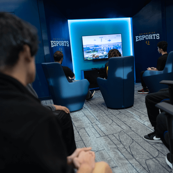 3 students sit in lounge chairs in a blue-lit esports room, watching and playing a competitive video game on a large wall-mounted screen, with Quinnipiac Esports branding visible on the walls.