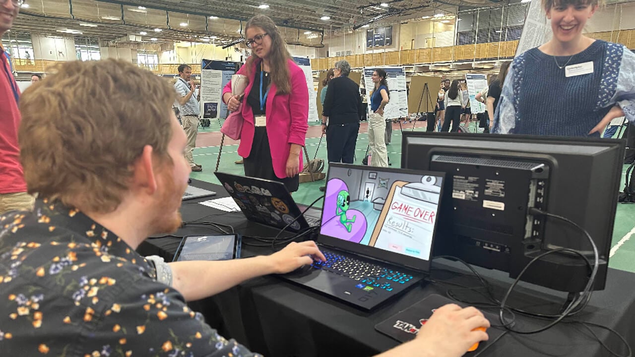 A faculty member in a patterned shirt tries out a game on a laptop. The screen shows a cartoon green alien in a purple chair and the words "Game Over."